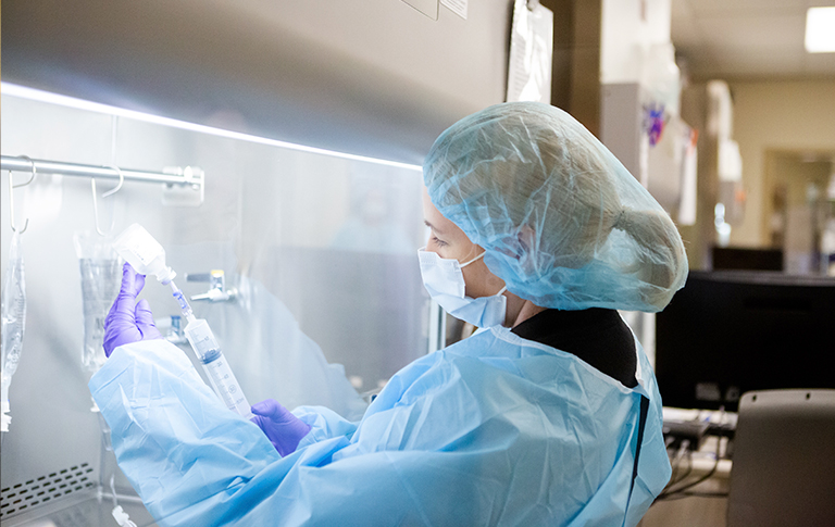 Gowned professional in a lab using a syringe and IV bag.